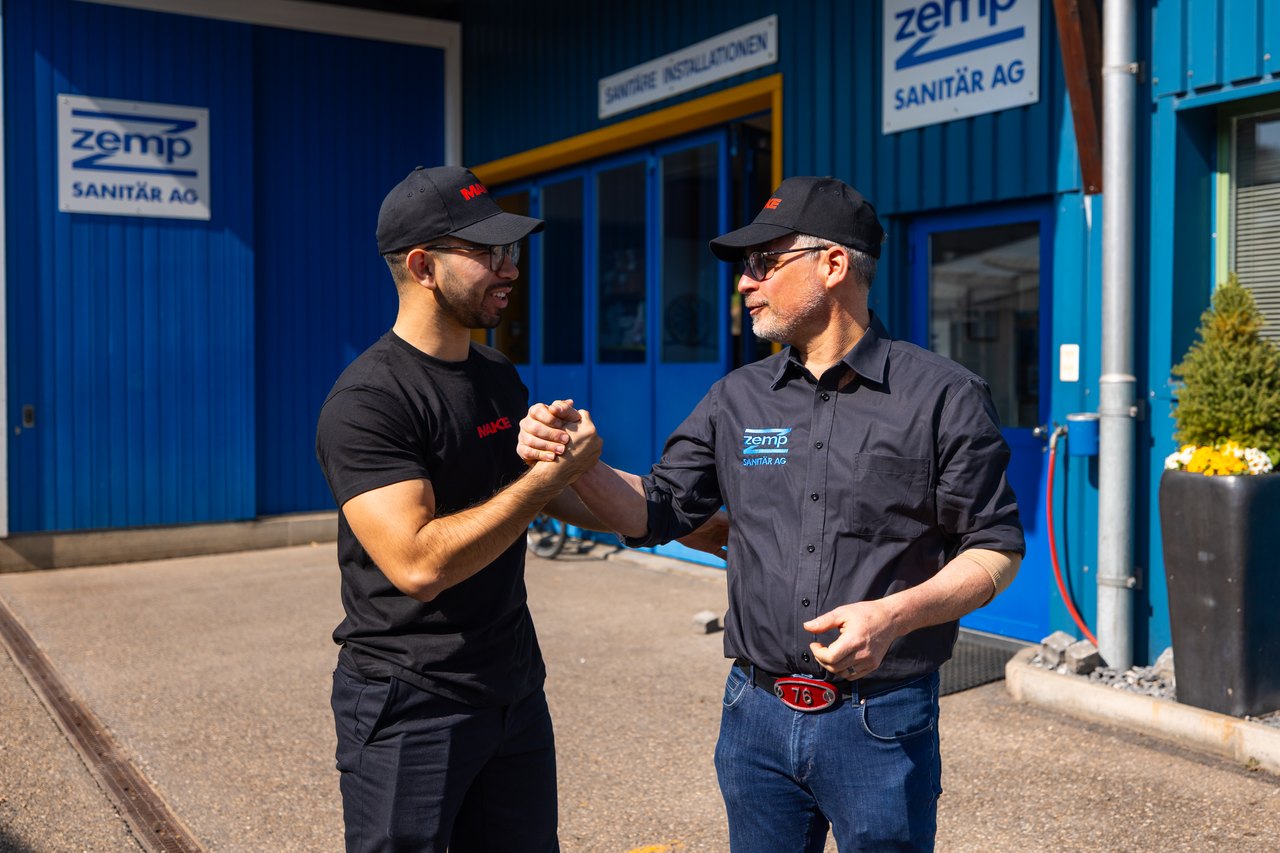 Zwei Männer in schwarzen Shirts und Kappen schütteln sich die Hände vor einem blauen Gebäude mit "zemp SANITÄR AG" Schriftzug.