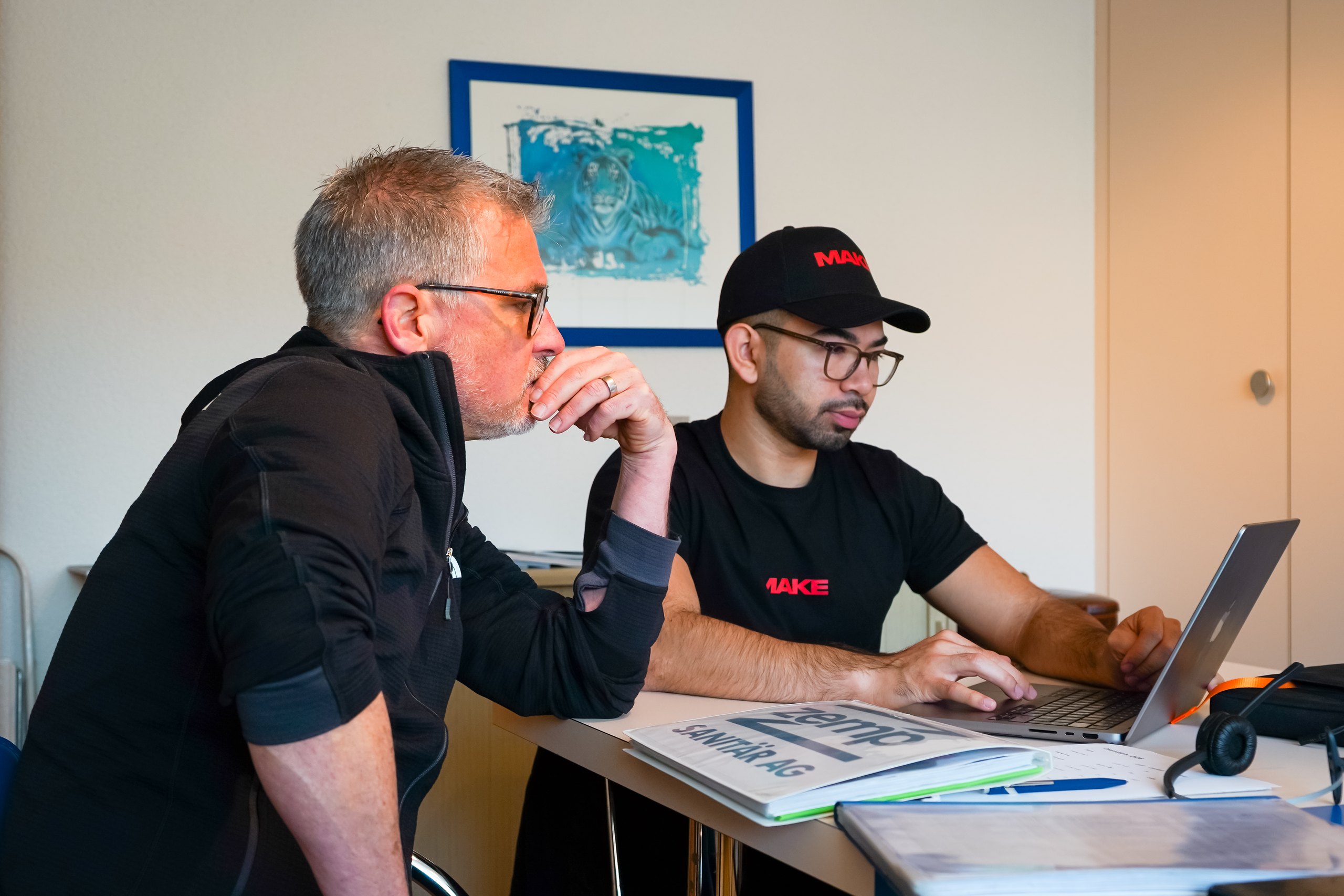 Zwei Männer schauen auf einen Laptop, der jüngere tippt, der ältere denkt nach.