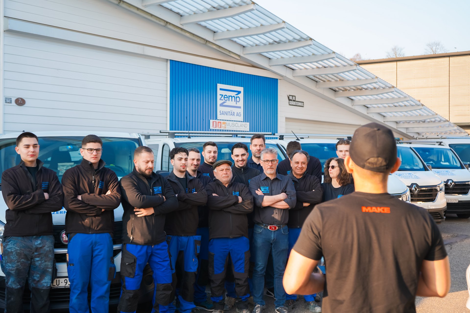 Eine Gruppe von 14 Männern und einer Frau in blauen Arbeitsanzügen steht vor weißen Transportern und einem Gebäude mit „zemp“-Logo.