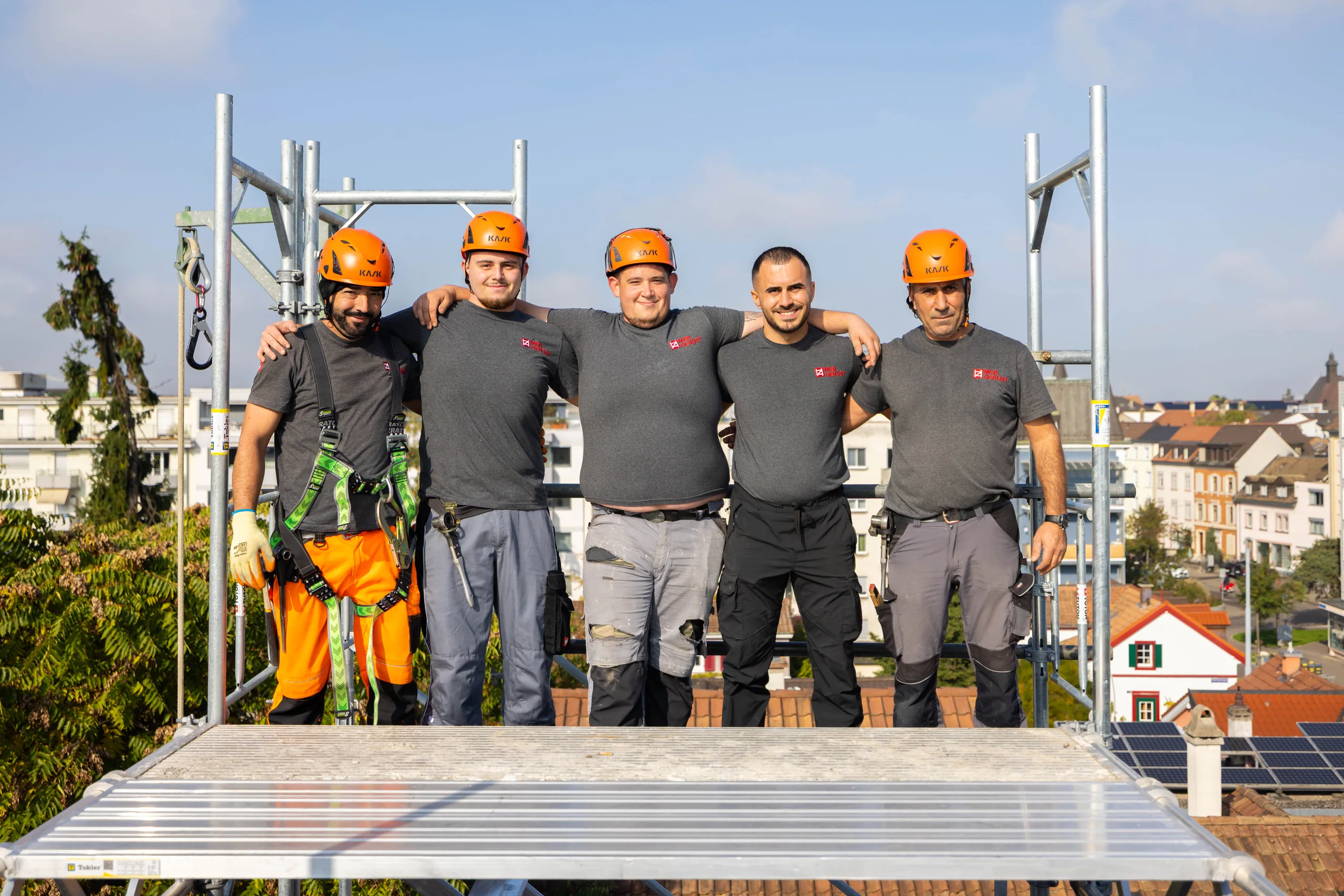 Fünf Gerüstbauer von MEB Gerüst mit orangen Helmen auf einer Gerüstplattform über den Dächern von Basel